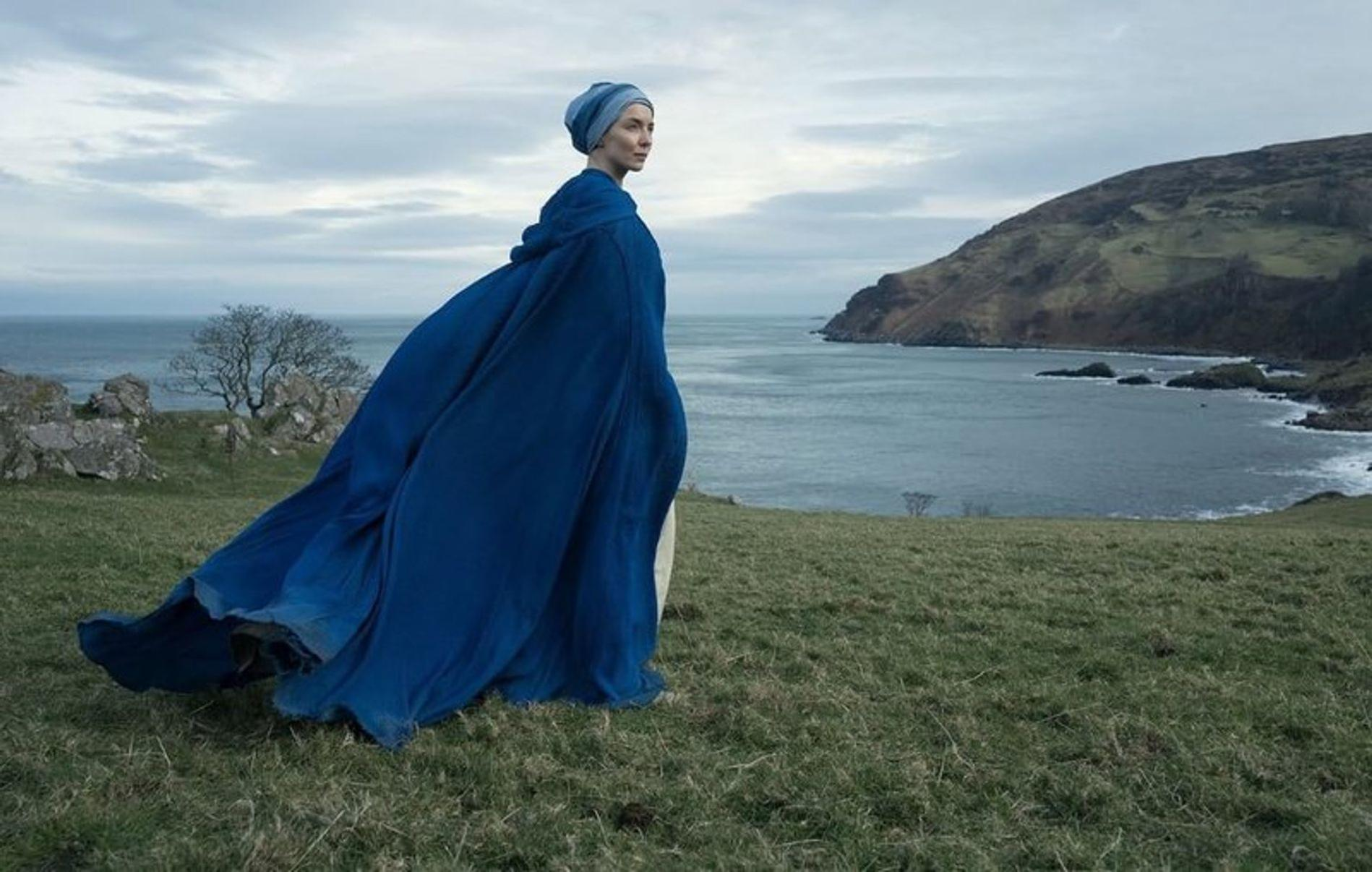 Frau in blauem Umhang blickt aufs Meer, Küste im Hintergrund.