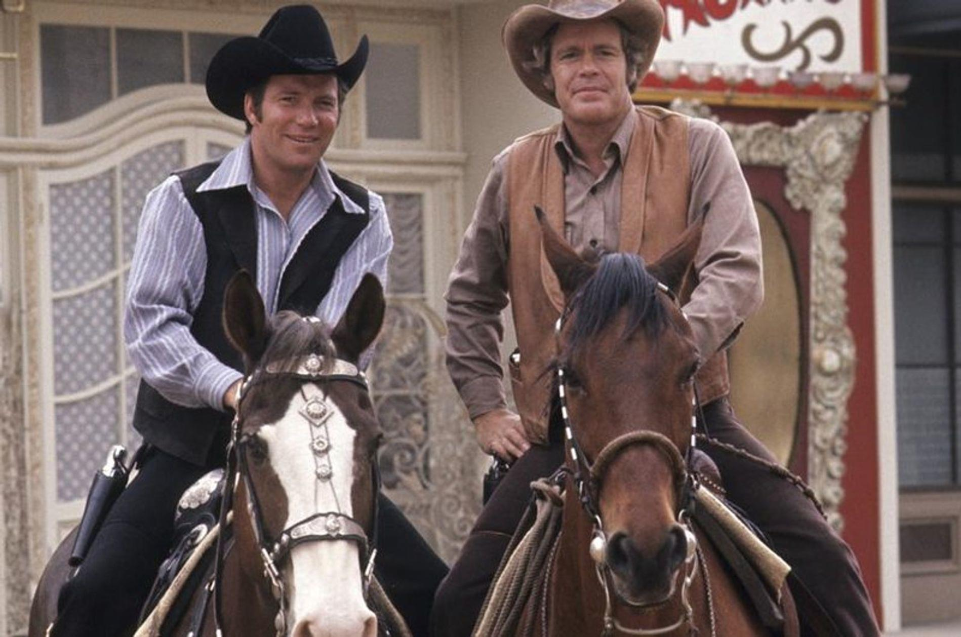 Zwei Männer in Cowboy-Kleidung reiten auf Pferden vor einem Gebäude.