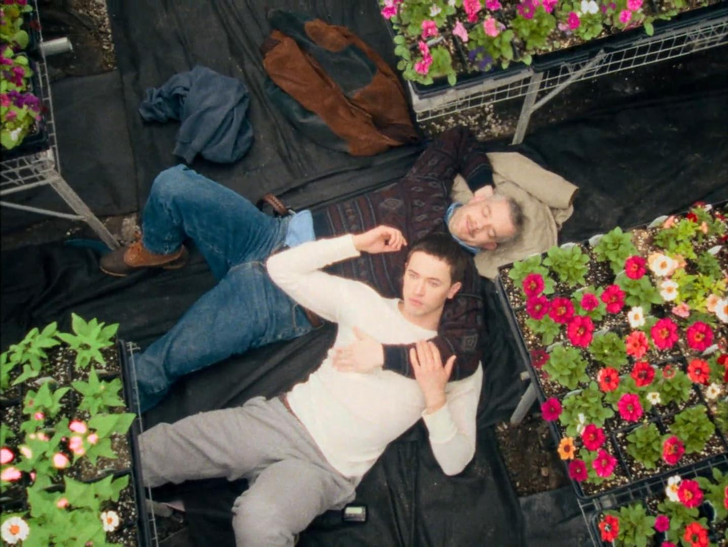 Zwei Männer liegen im Arm, umgeben von Blumen in einem Gewächshaus.