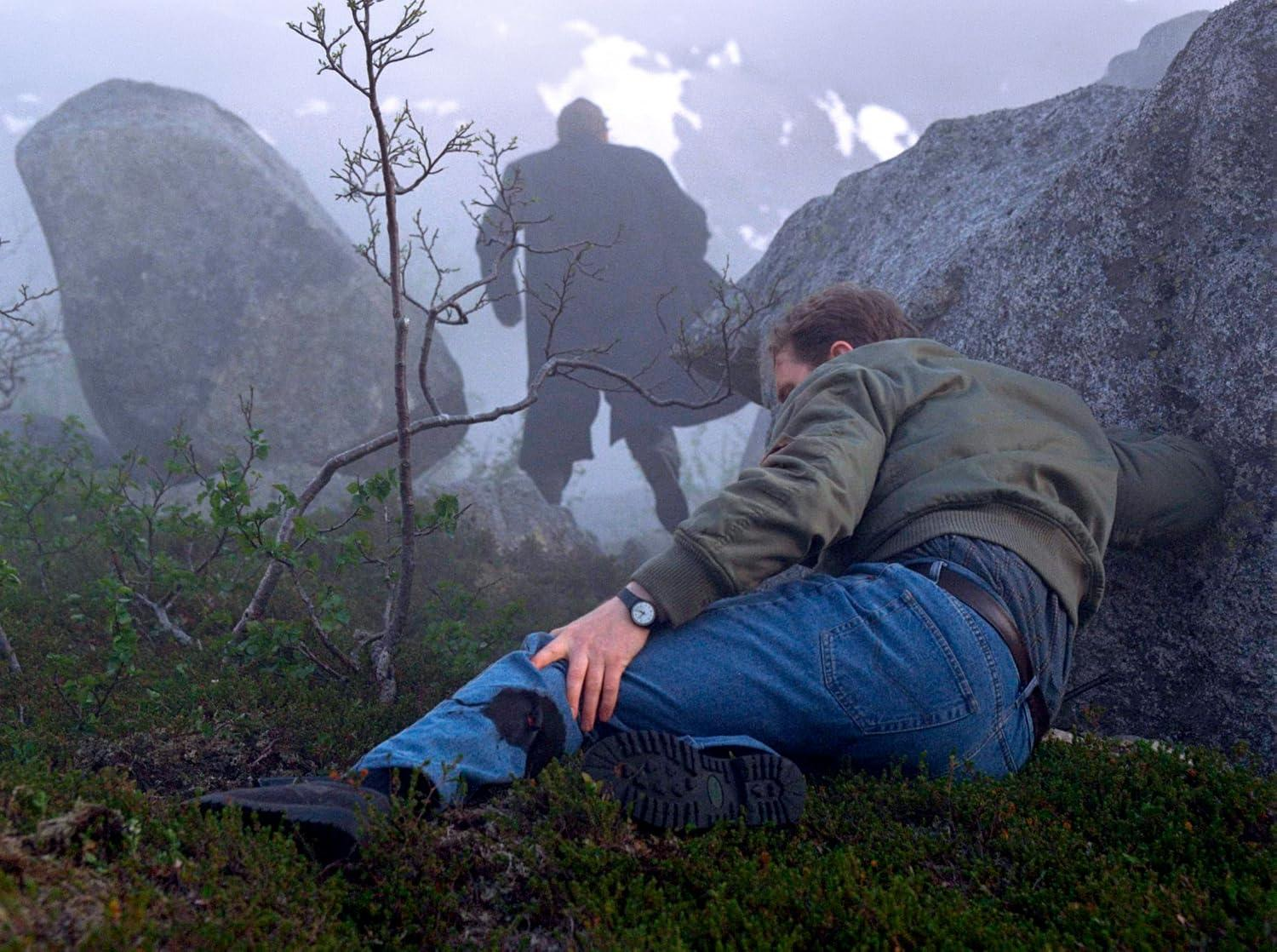 Mann sitzt auf moosigem Boden. Ein Mann im Mantel steht im Nebel hinter ihm.