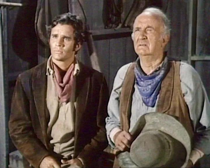 Zwei Männer in westlicher Kleidung. Der Jüngere trägt eine braune Jacke und einen roten Schal. Der Ältere trägt eine Weste und ein blaues Bandana.