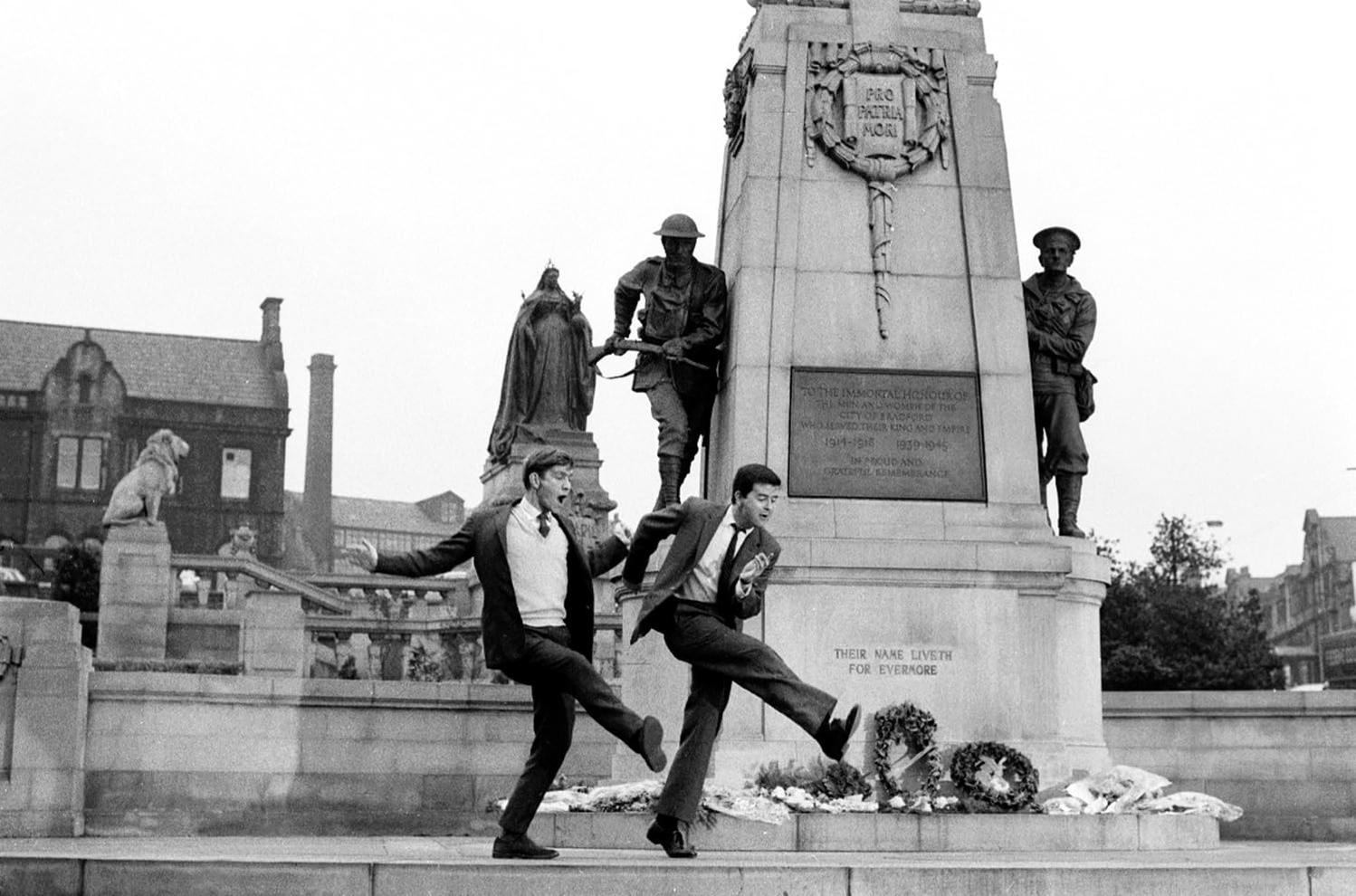Zwei junge Männer tanzen vor einem Kriegerdenkmal mit Soldatenstatuen.
