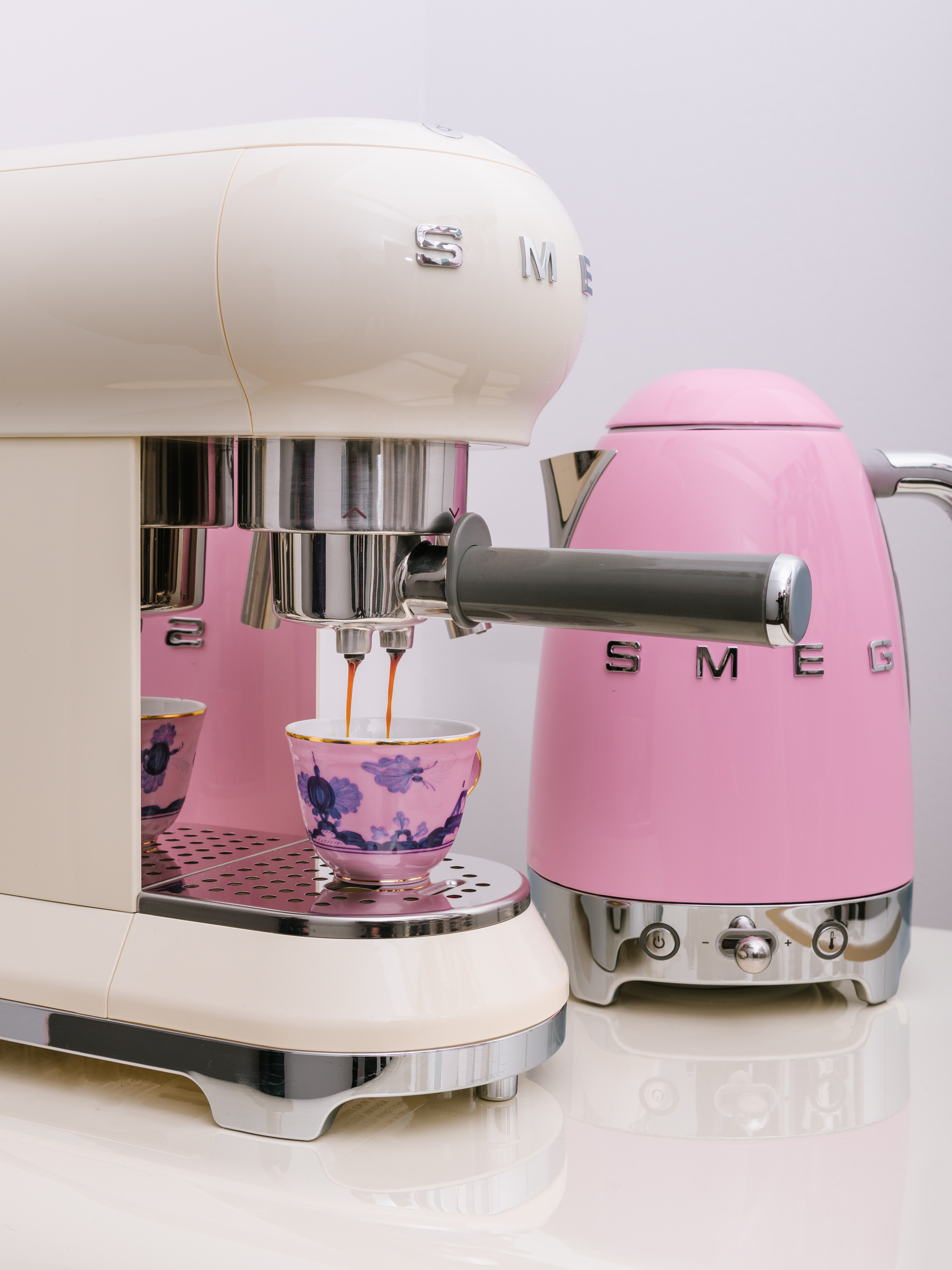 Eine cremefarbene Espressomaschine brüht Kaffee in eine rosa Tasse. Daneben steht ein pinker Wasserkocher.