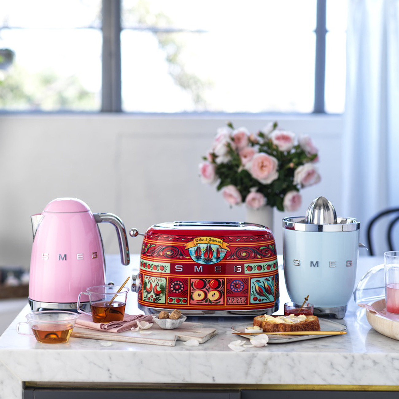 Rosa Wasserkocher, roter Toaster und blauer Entsafter auf Marmortheke mit Tee und Toast.