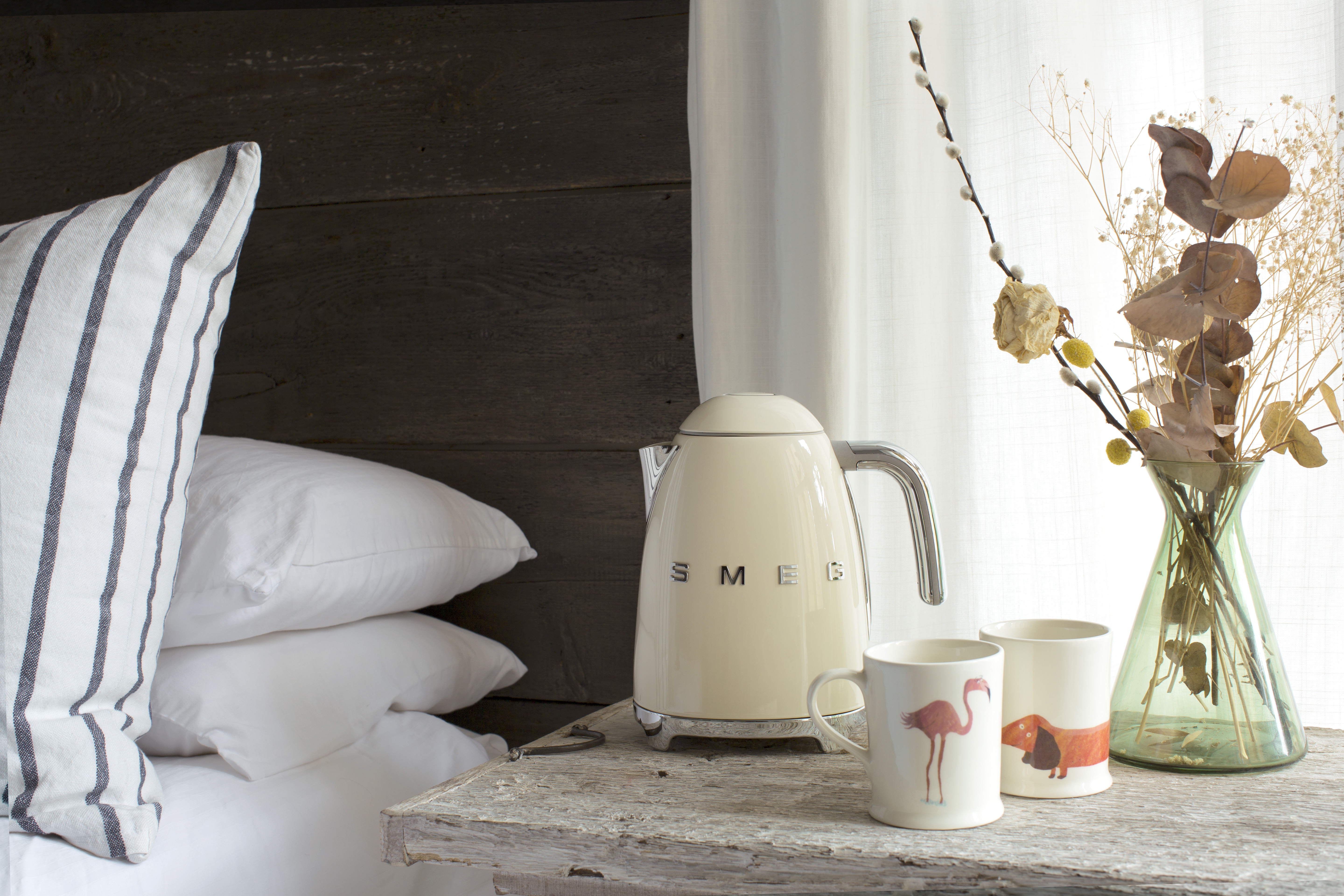 Ein cremefarbener Smeg-Wasserkocher steht auf einem Holztisch neben zwei Tassen mit Tiermotiven und einer Vase mit Trockenblumen.