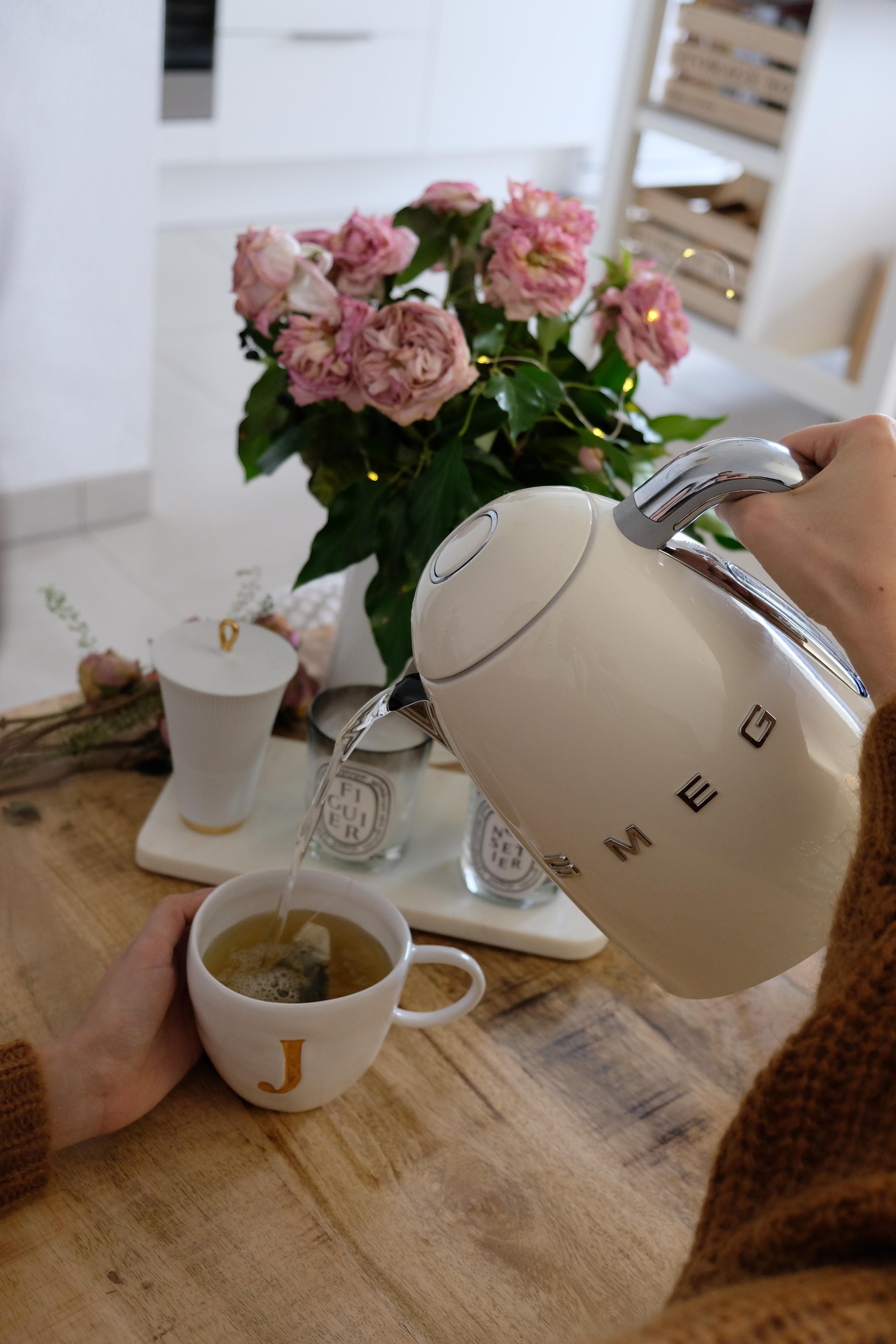 Eine weiße Smeg-Kanne gießt heißes Wasser in eine Teetasse mit dem Buchstaben J.