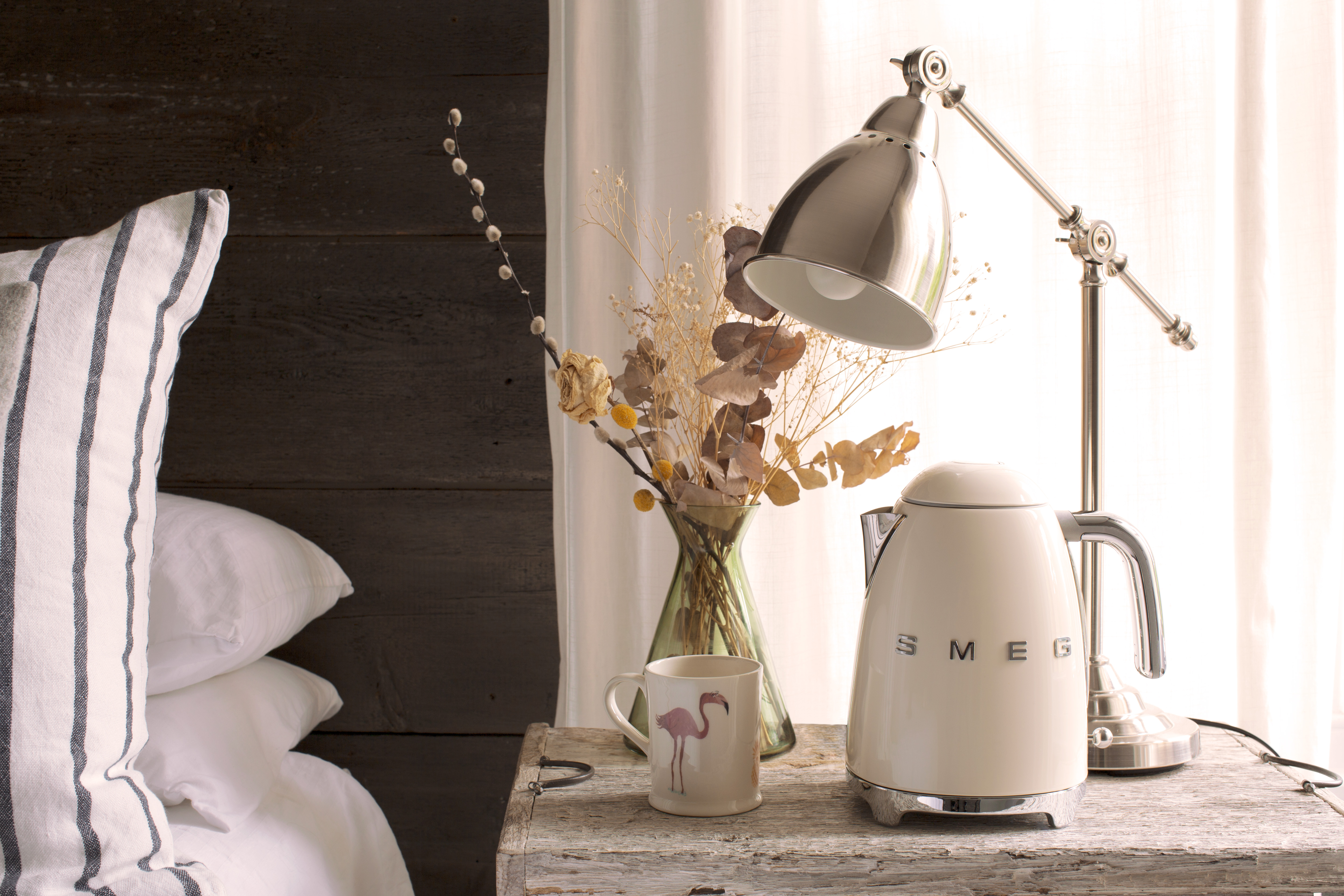 Ein cremefarbener Wasserkocher, eine Tasse mit Flamingos und eine Lampe auf einem Holztisch.