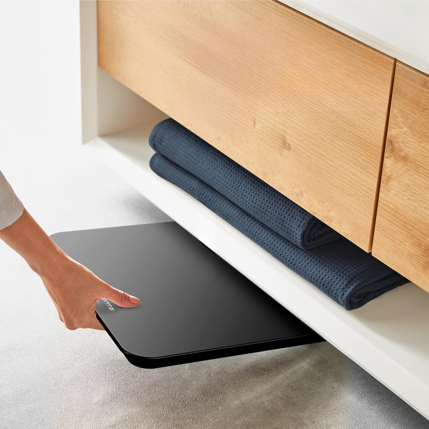 Eine Hand zieht eine schwarze Waage unter einem Holzschrank hervor.