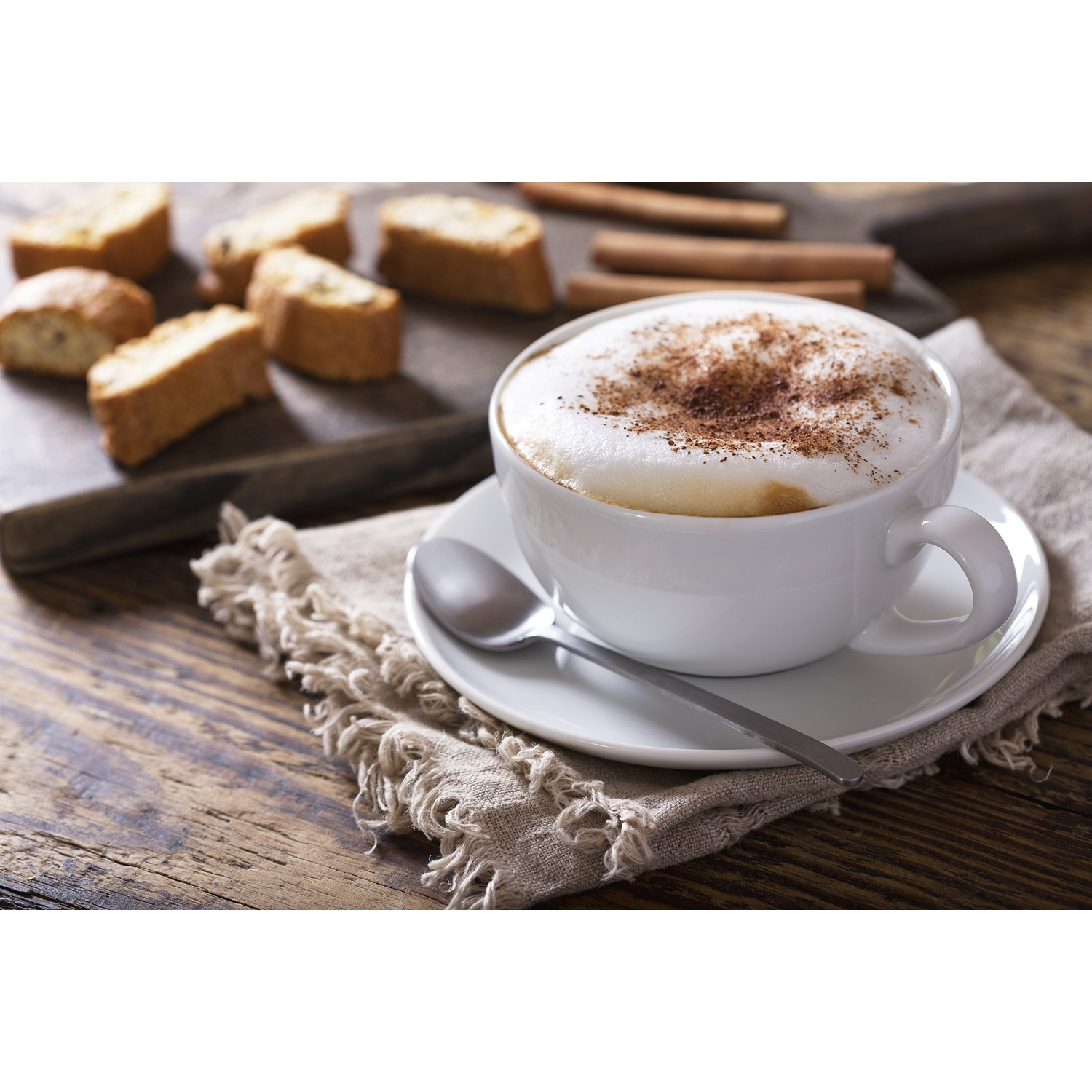 Una tazza di cappuccino con schiuma e cannella accanto a cantuccini e bastoncini di cannella.