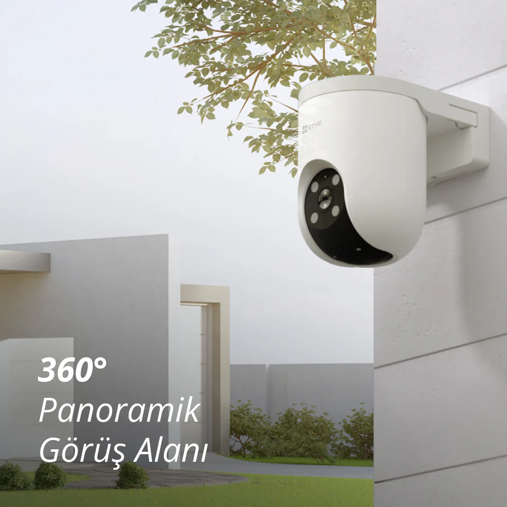 Beyaz güvenlik kamerası ev duvarına monte edilmiş, arkada ağaç ve bina var.