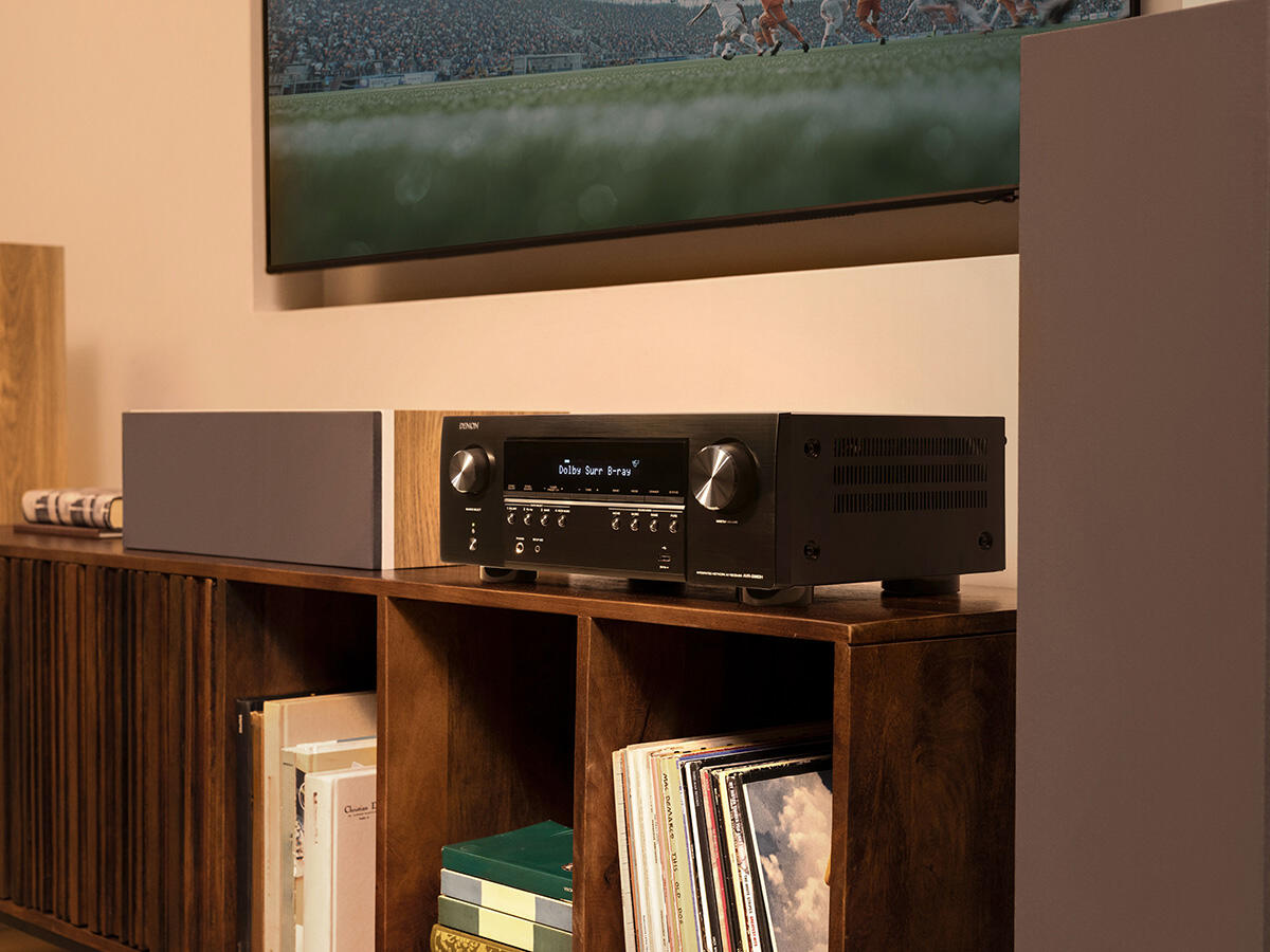 Egy fekete Denon AV-vevő áll egy fa polcon egy televízió alatt, amely focimeccset mutat.