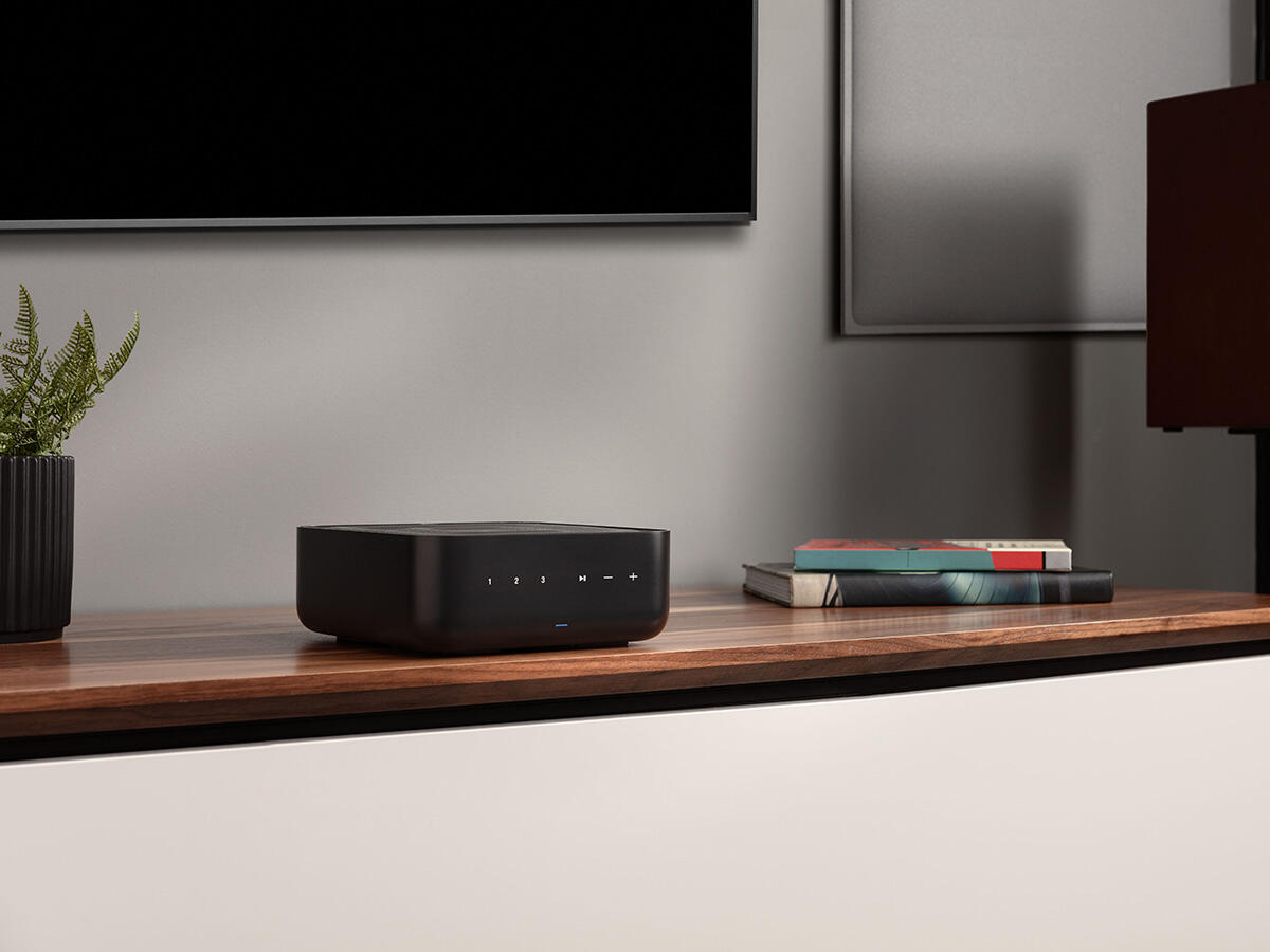 Egy fekete audiostreamer áll egy fa szekrényen, egy növényes cserép és könyvek mellett.