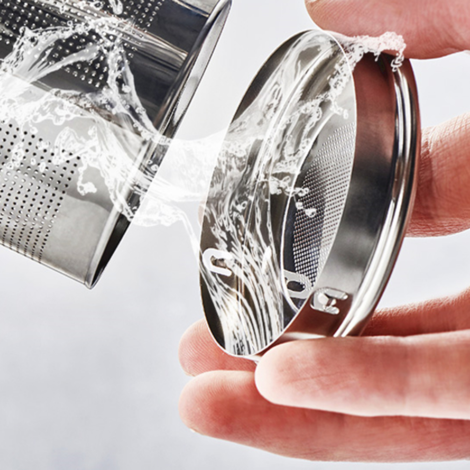 Een hand houdt een zilveren theefilter vast waar water doorheen stroomt.