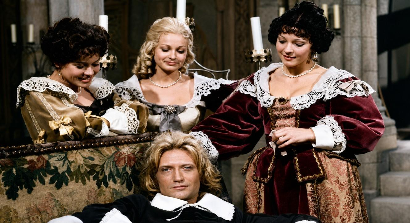 Ein Mann mit blondem Haar liegt auf einem Sofa, umgeben von drei Frauen in historischen Kleidern.