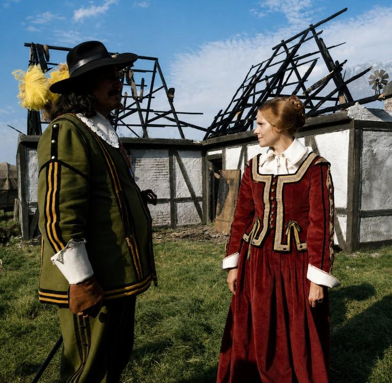 Ein Mann in historischer Kleidung und eine Frau stehen vor einem ausgebrannten Gebäude.