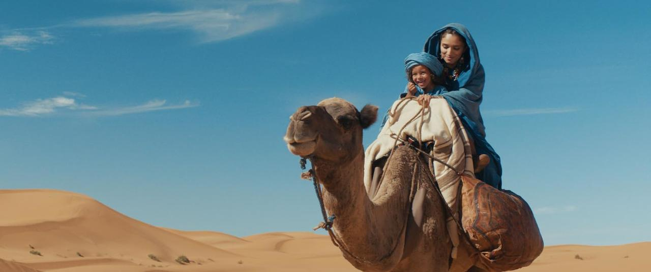 Eine Frau und ein Kind reiten auf einem Kamel durch Sanddünen unter blauem Himmel.