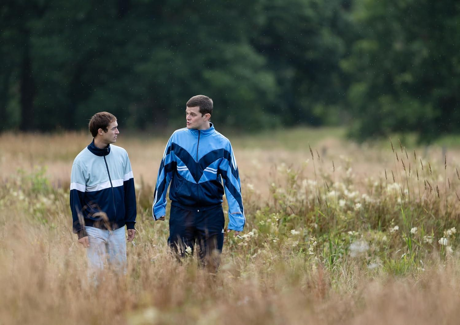 Zwei Männer in Trainingsanzügen gehen durch hohes Gras, Bäume im Hintergrund.