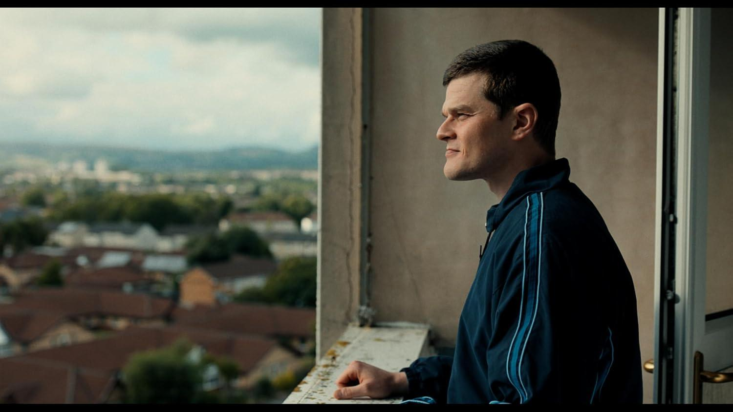 Ein Mann in einem blauen Trainingsanzug steht auf einem Balkon und blickt auf eine Stadt.