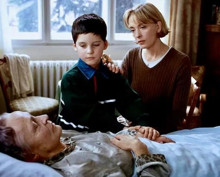 Ein Junge und eine Frau blicken auf eine ältere Person im Bett. Die Frau legt die Hand auf die Schulter des Jungen.