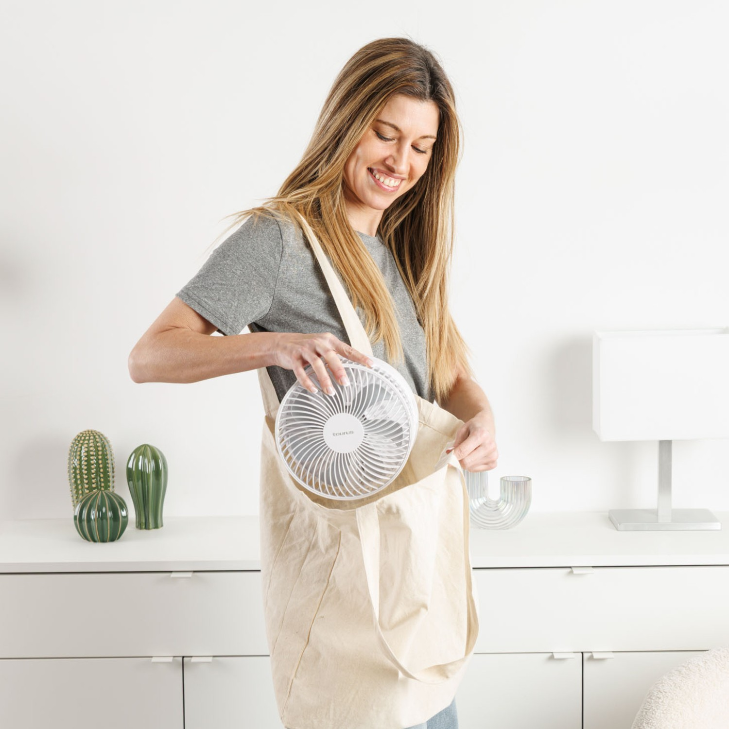 Una mujer mete un ventilador pequeño en una bolsa de tela. Jarrones de cactus están en una cómoda.