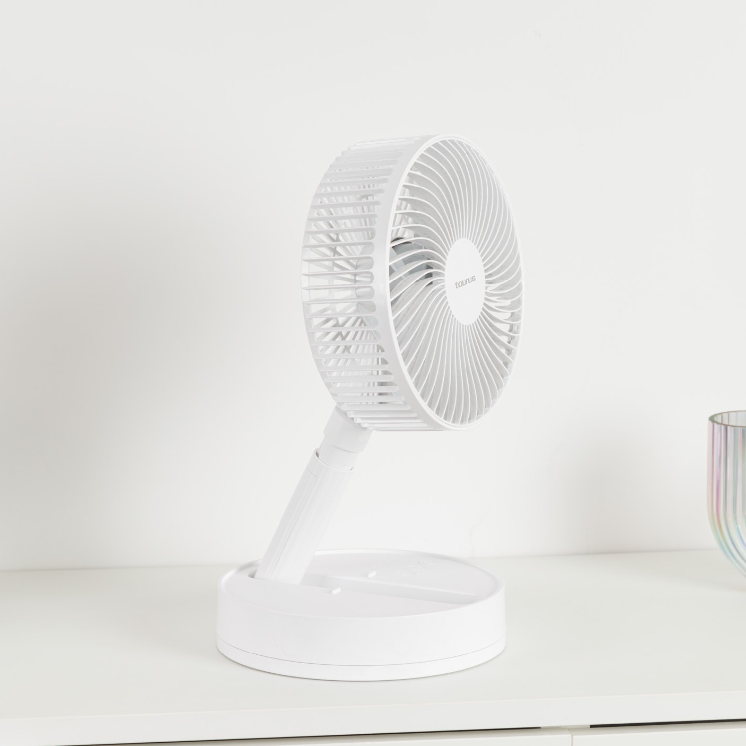 Un ventilador blanco portátil está sobre una mesa blanca. A la derecha hay un jarrón de cristal.