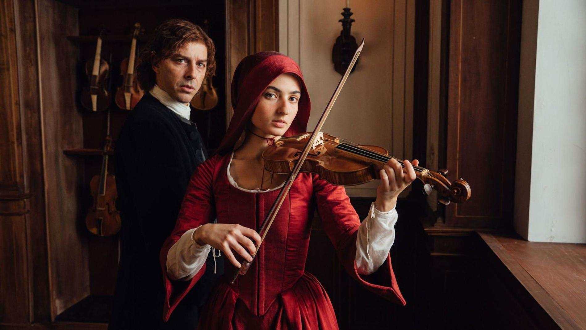 Frau in Rot spielt Violine, Mann im schwarzen Mantel steht dahinter.