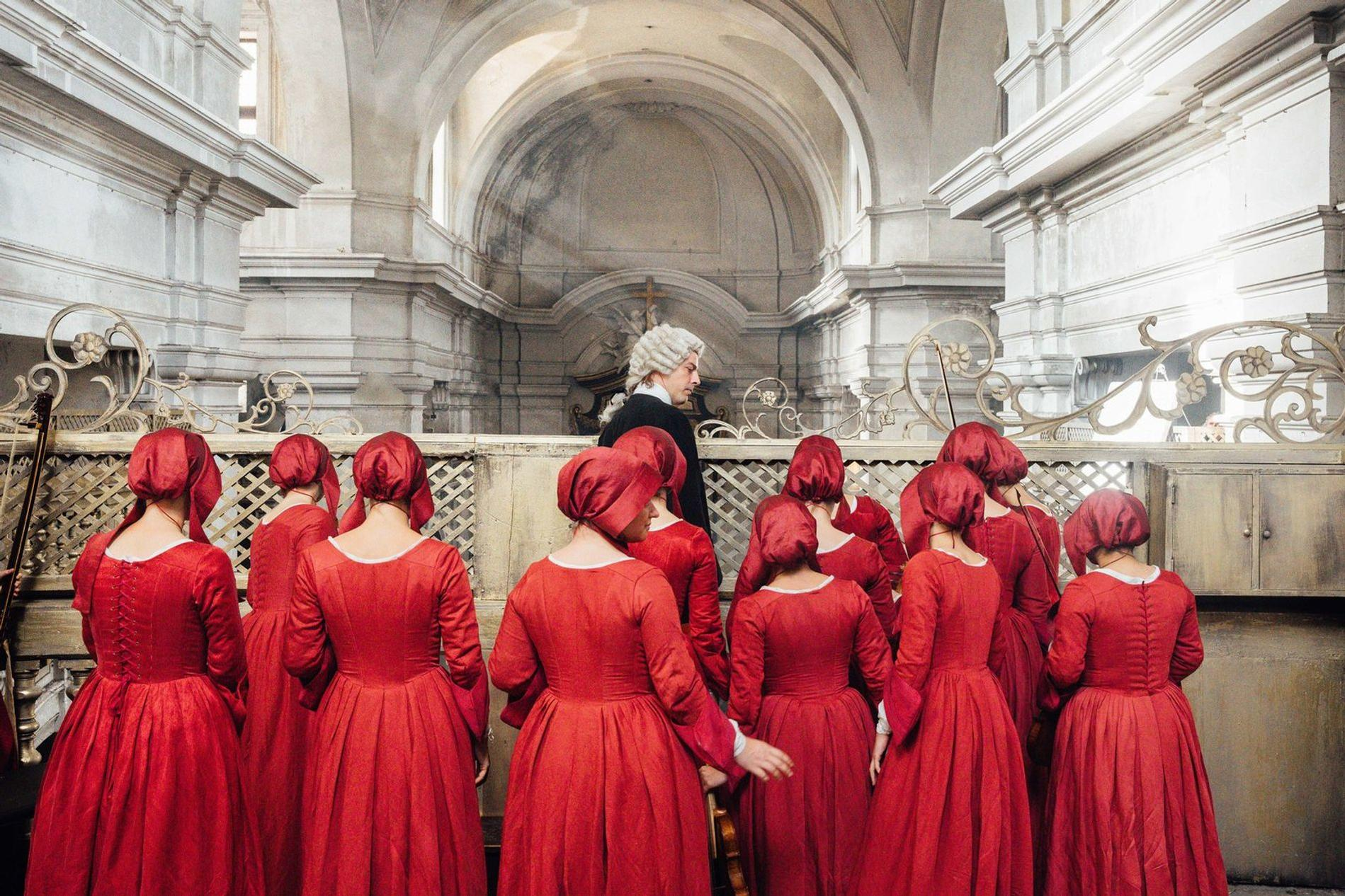 Männer in Perücke und Frauen in roten Kleidern stehen in einer Kirche.
