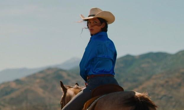 Eine Frau in einem Cowboyhut reitet auf einem Pferd vor hügeliger Landschaft.