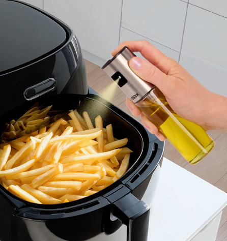 Une main pulvérise de l'huile d'un vaporisateur en verre sur des frites dans une friteuse à air noire.