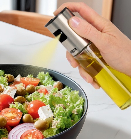 Un vaporisateur d'huile d'olive est pulvérisé sur une salade avec tomates, olives et feta.