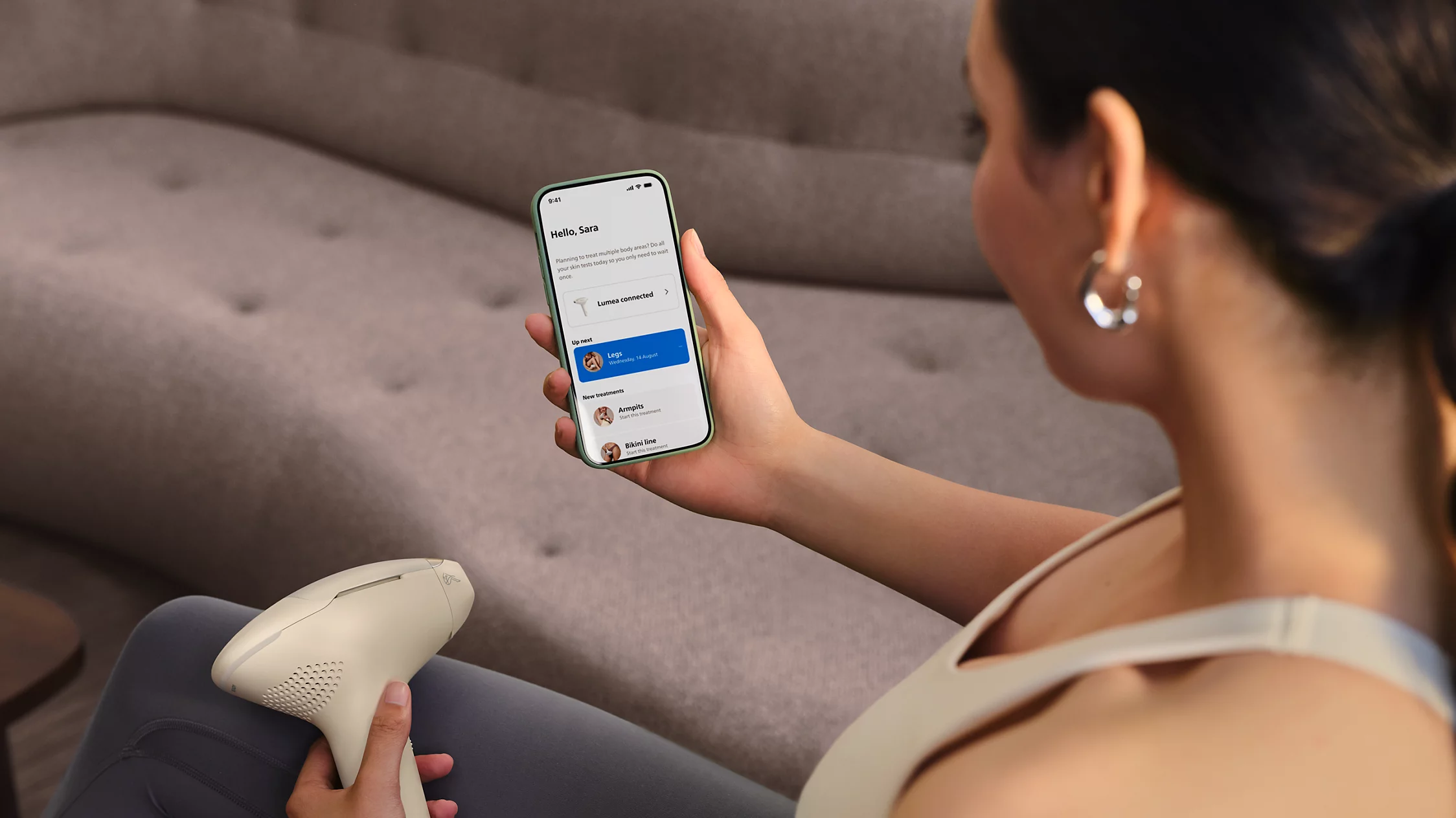 Frau hält IPL-Gerät und Handy mit Lumea-App. Beine-Behandlung geplant.