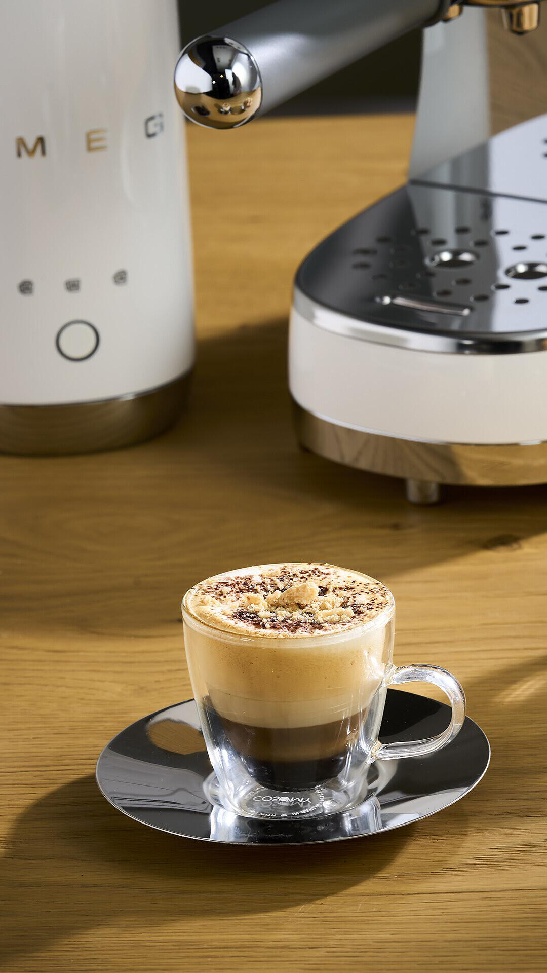 Ein Glas Kaffee mit Schichten steht auf einer silbernen Untertasse, im Hintergrund eine weiße Kaffeemaschine.