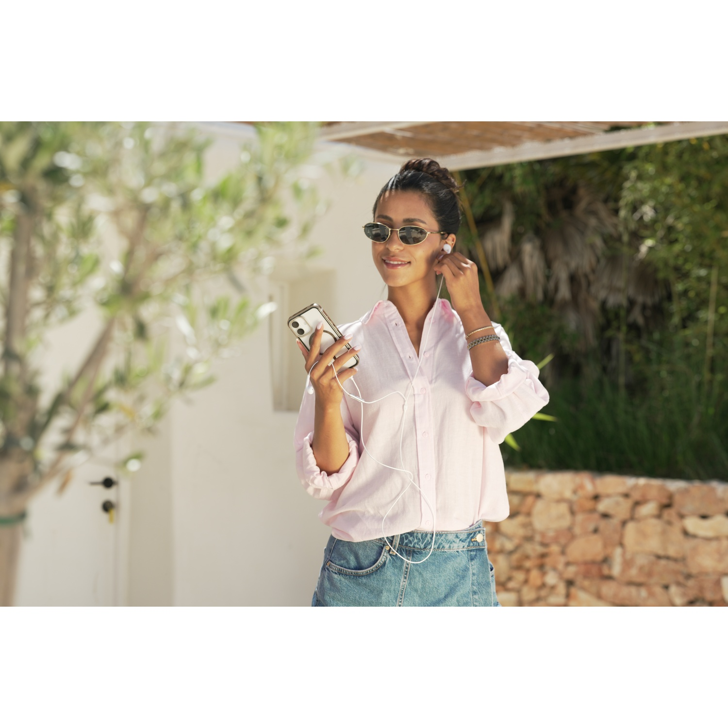 Una donna con occhiali da sole e auricolari tiene in mano uno smartphone.