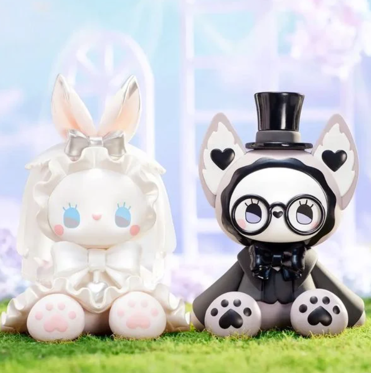 Zwei Figuren auf Gras: eine weiße Hasenfigur mit Schleife und eine graue Katzenfigur mit Zylinder.