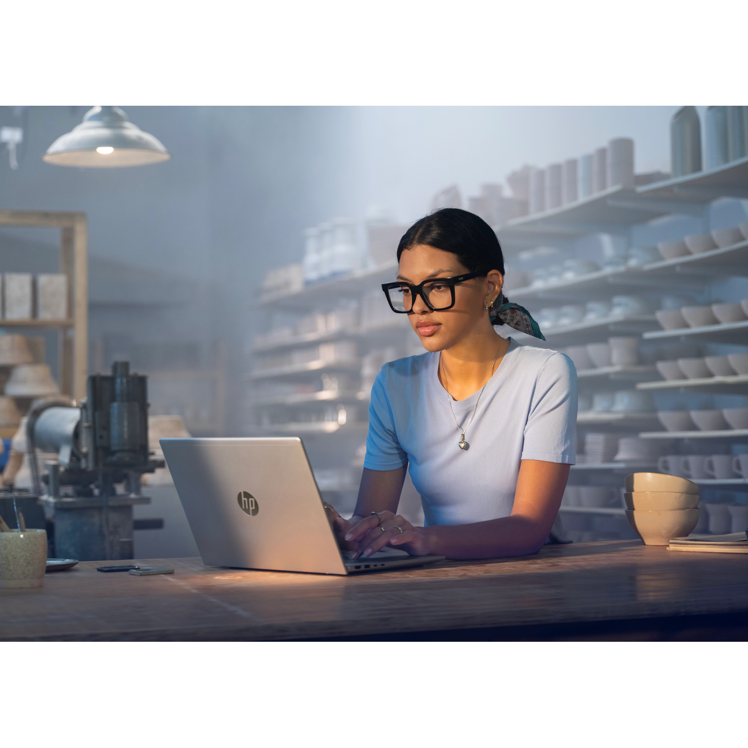 Een vrouw met bril typt op een laptop in een pottenbakkerij met keramiek op planken.