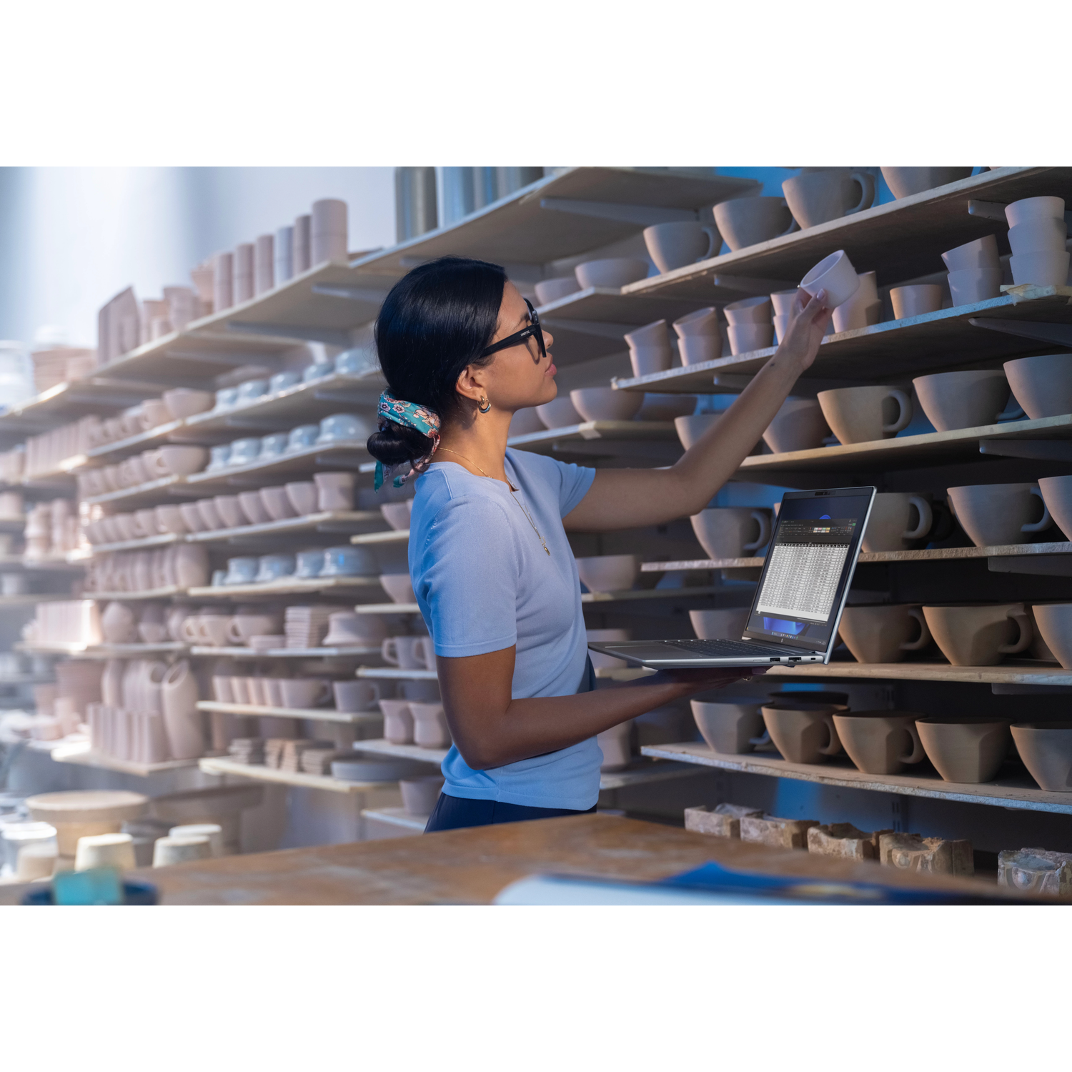 Een vrouw in een blauw shirt sorteert kopjes op planken en houdt een laptop vast.