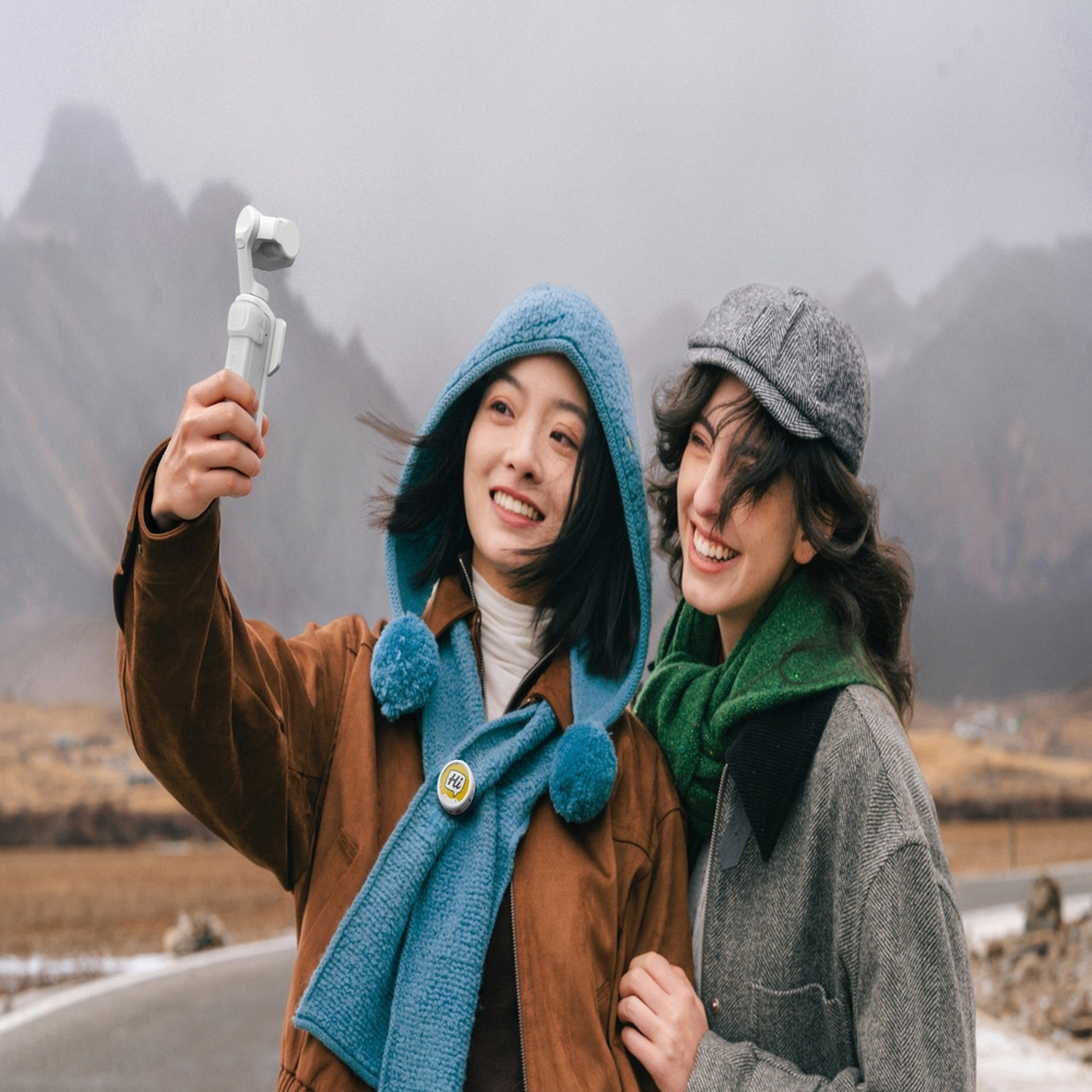 Due donne sorridenti in cappotti invernali si scattano un selfie con un gimbal in un paesaggio montano nebbioso.