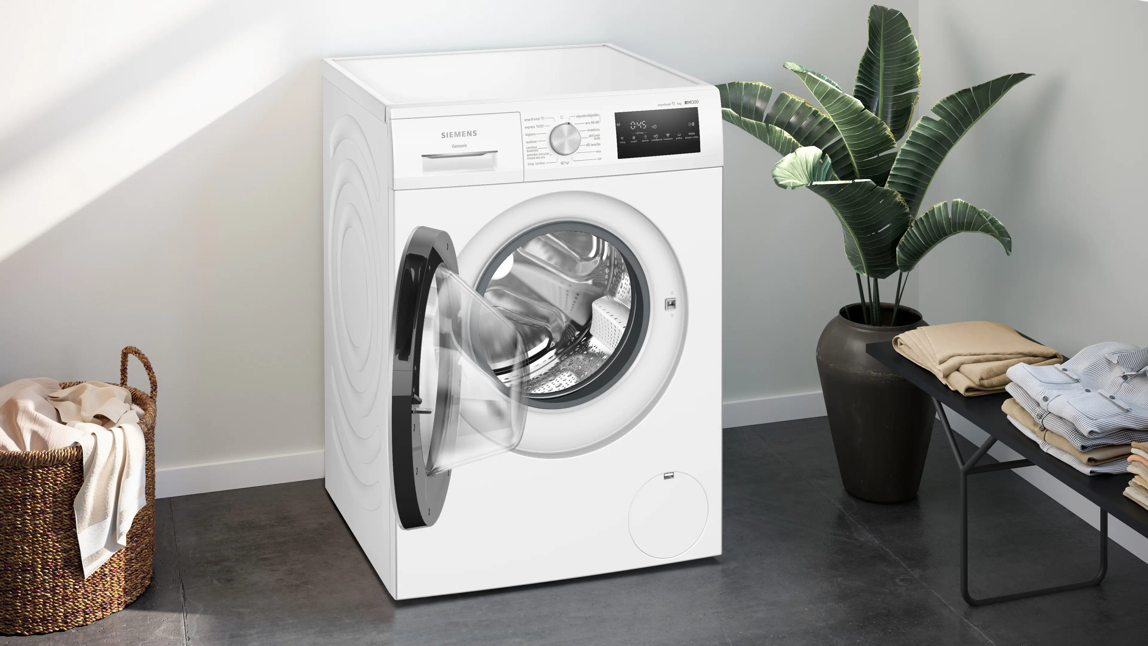 Una lavadora Siemens blanca con la puerta abierta, una cesta de ropa al lado.