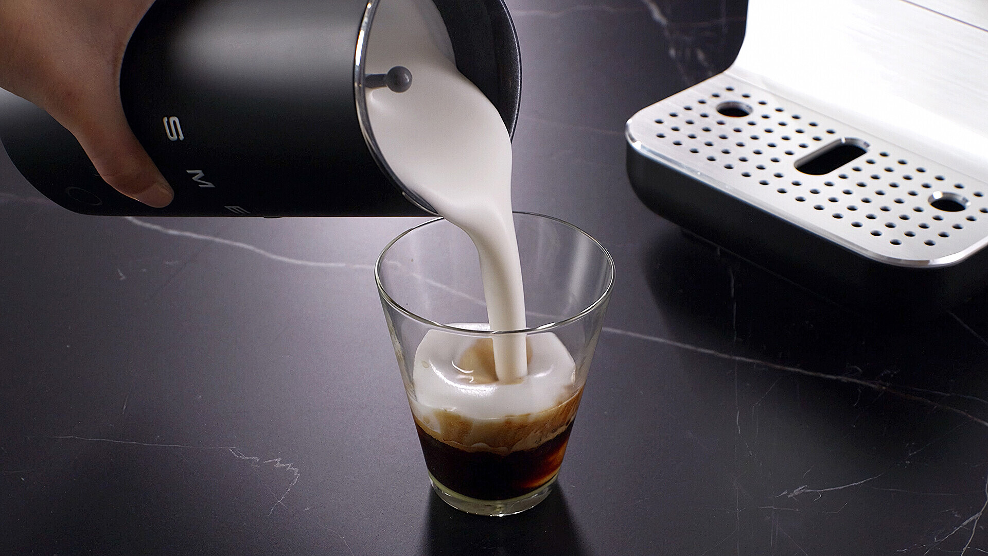 Milch wird in ein Glas mit Kaffee gegossen, ein Milchaufschäumer ist daneben.