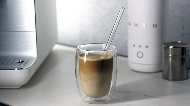 Ein Glas Latte Macchiato mit Strohhalm steht neben einem weißen Smeg-Gerät.