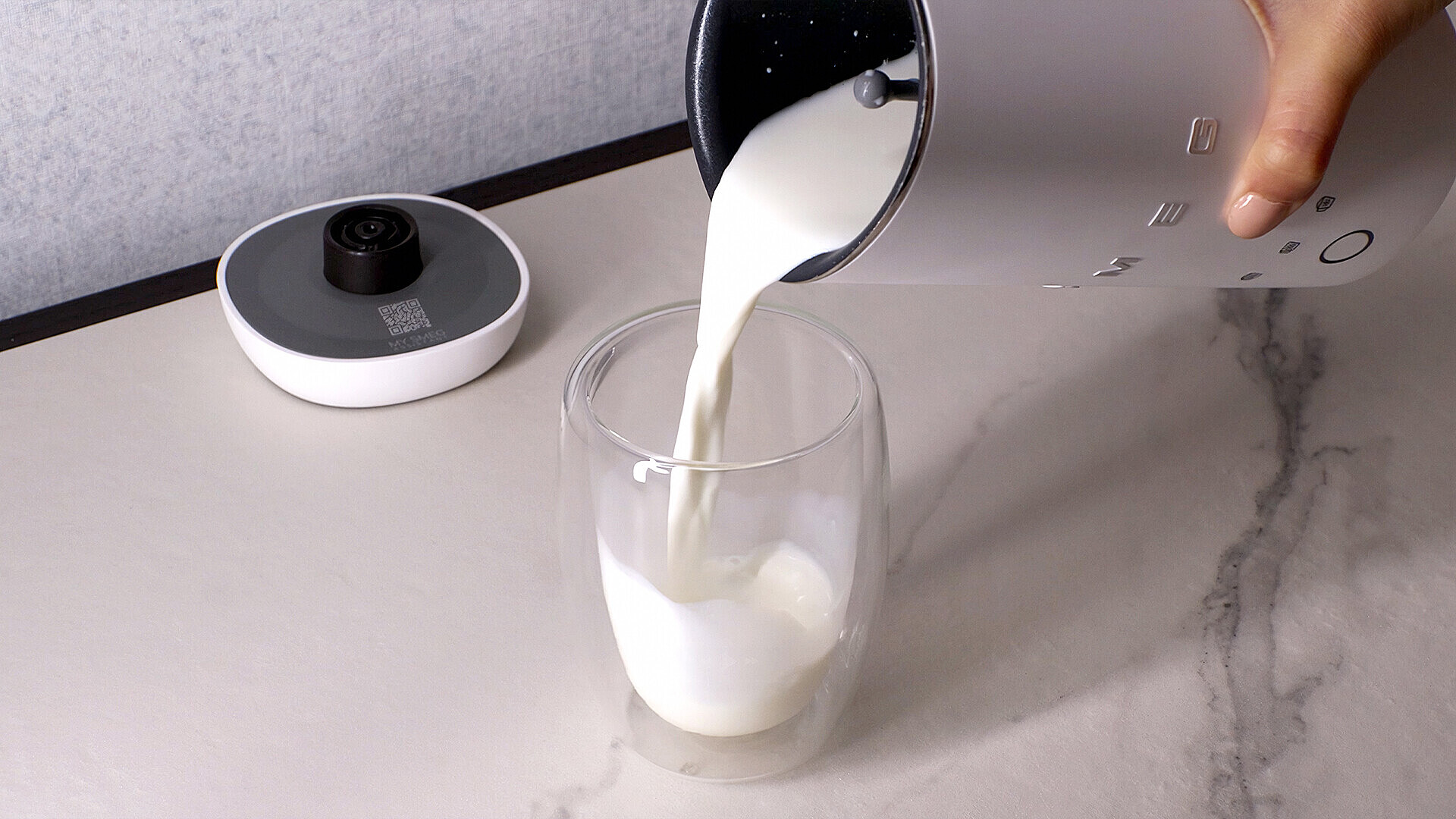 Milch wird aus einem silbernen Krug in ein klares Glas gegossen. Links steht ein Gerät.
