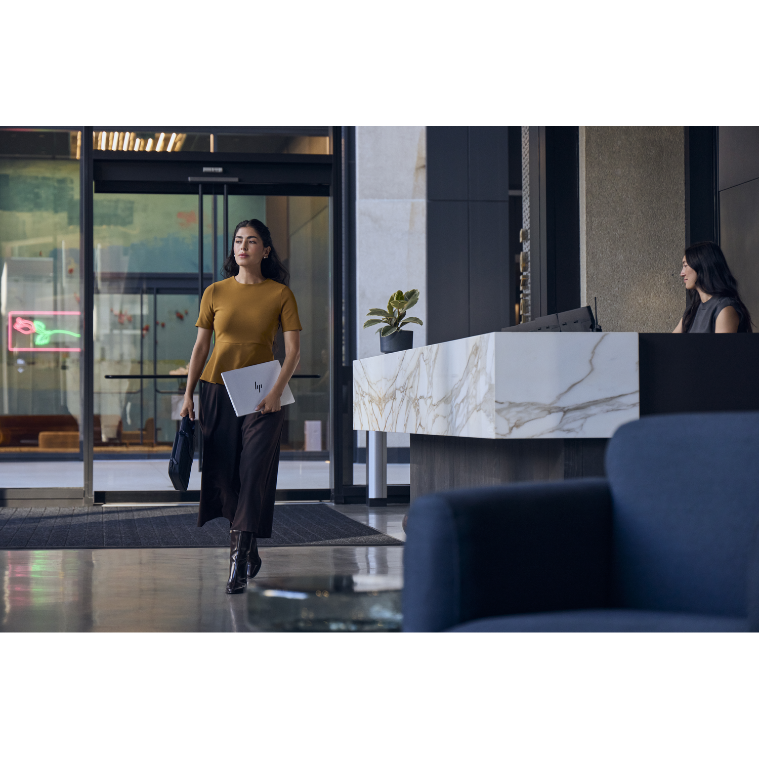 Een vrouw met een laptop loopt door een lobby, een receptioniste is op de achtergrond.
