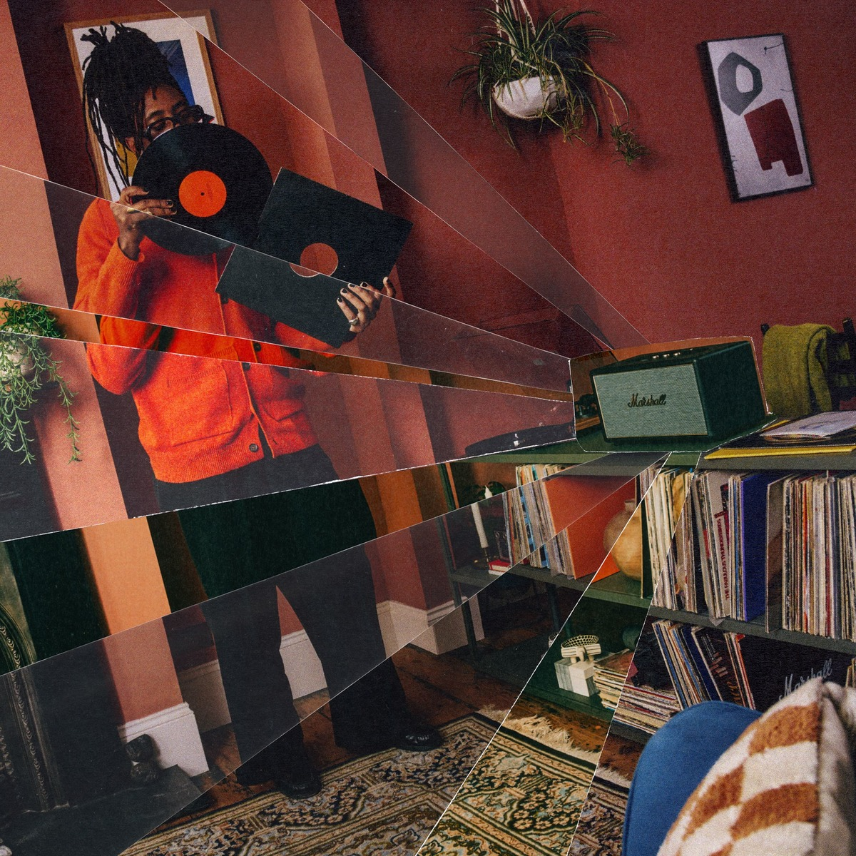 Eine Person in einem orangen Pullover hält Schallplatten vor einem Regal mit Schallplatten und einem Lautsprecher.
