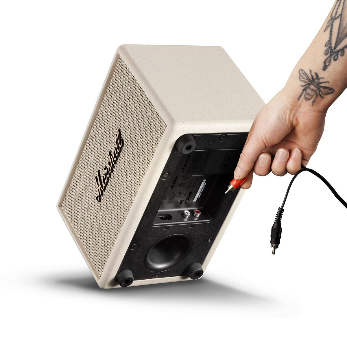 Eine Hand mit einem Bienen-Tattoo verbindet ein rotes Cinch-Kabel mit einem beigen Marshall-Lautsprecher.