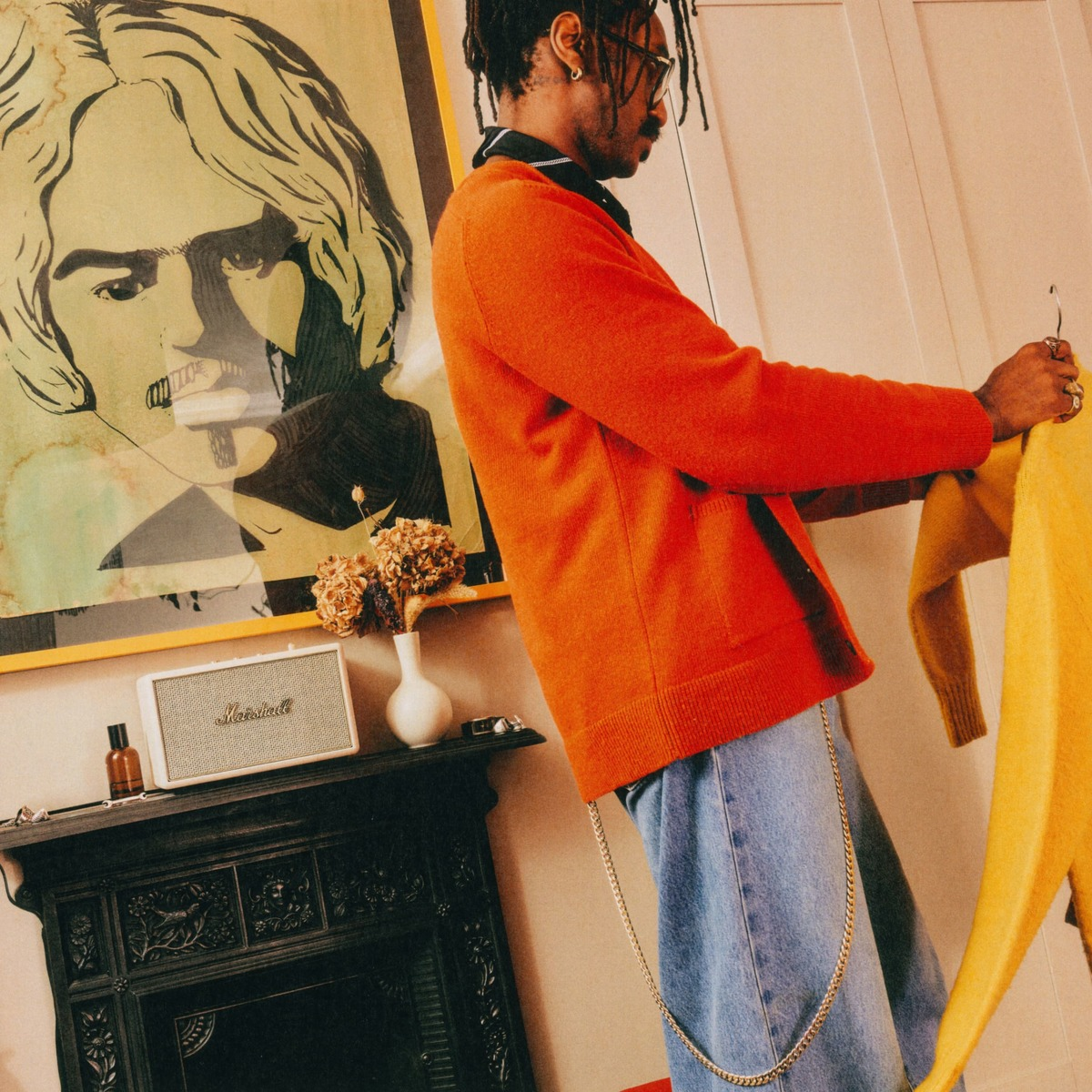 Ein Mann im orangen Pullover und Jeans hält ein gelbes Kleidungsstück vor einem Kamin mit Kunst.