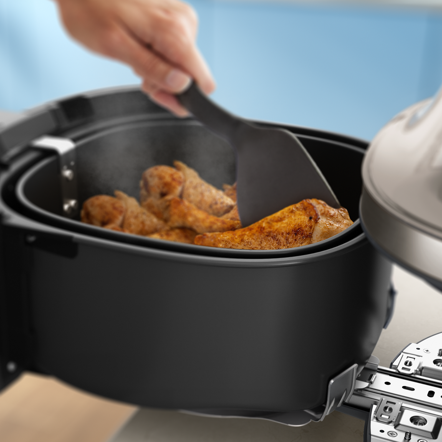 Une main utilise une spatule pour retourner des pilons de poulet dorés dans une friteuse à air noire fumante.