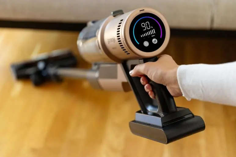 Egy kéz arany színű akkus porszívót tart digitális kijelzővel fa padlón.