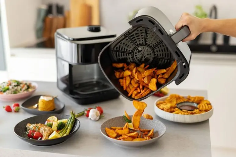 Egy air fryer sült krumplit önt egy tálba. Közelben csirkés és zöldséges ételek vannak.
