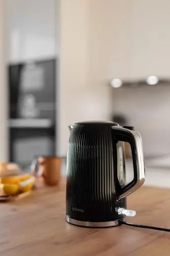 Egy fekete, bordázott vízforraló áll egy faasztalon. Konyha a háttérben.