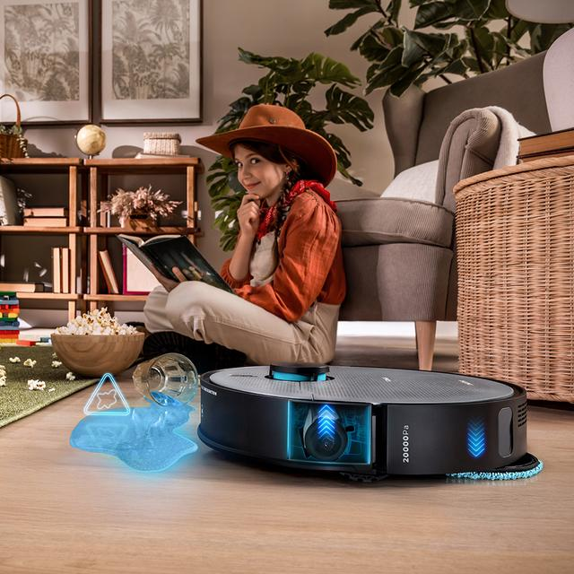 Şapkalı bir kız, robot süpürge su dökülmesini temizlerken kitap okuyor.